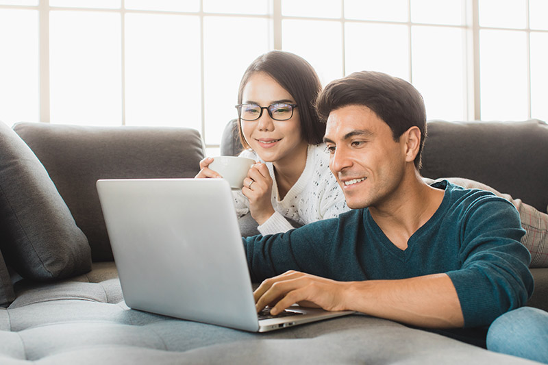 A smiling couple sits on a couch, looking at a laptop together. The woman holds a white mug, and both appear relaxed and engaged. Large windows in the background let in natural light.