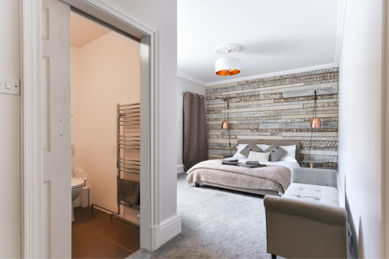 A modern bedroom with a rustic wood accent wall, double bed, neutral bedding, wall lamps, gray bench, and an open door leading to a small en-suite bathroom with a towel warmer.